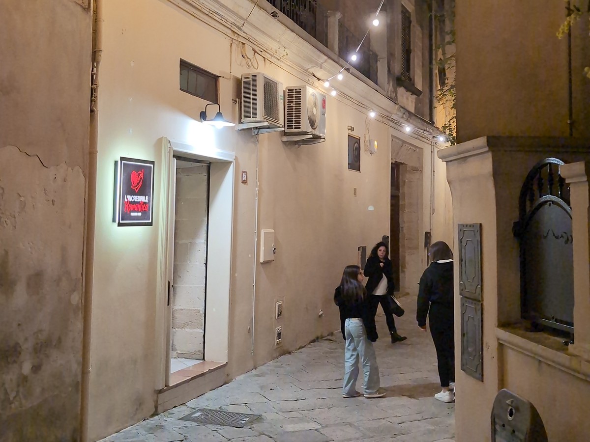 Ingresso serale de L'Incredibile Romantica con insegna luminosa a cuore rosso in un vicolo lastricato con luci sospese